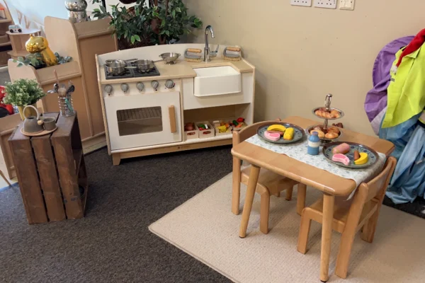 Wooden toy kitchenette and small wooden table set arranged with toy plates and play pastries for tea, placed on a rug in a children's play corner with crates, coats, and plants.