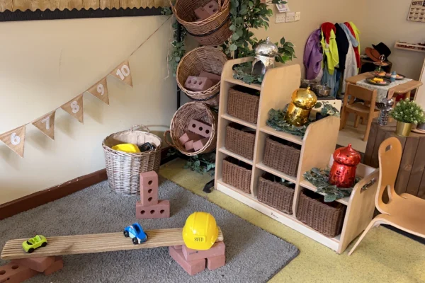 Yellow hard hat labeled "SMITH" rests on foam bricks beside a wooden seesaw with toy cars, in a preschool corner with baskets, dress-up coats, pennant 6 7 8 9 10.