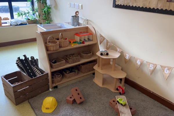 Wooden toy shelf holds baskets and play pieces while toy cars and foam bricks lie scattered; a yellow hard hat sits on a rug; numbered bunting "1 2 3 4 5 6" decorates a playroom corner.