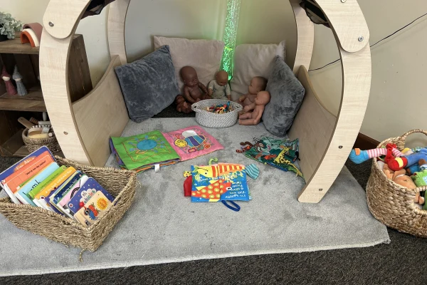 Wooden curved play pod cradles three baby dolls and a basket of beads; cloth and board books lie scattered on a soft gray carpeted mat in a small playroom corner.