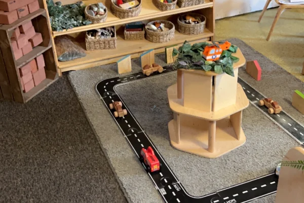 Red car drives on a black road mat around a low play tower topped by an orange helicopter; shelves, baskets, and blocks surround the carpeted area. No legible text.