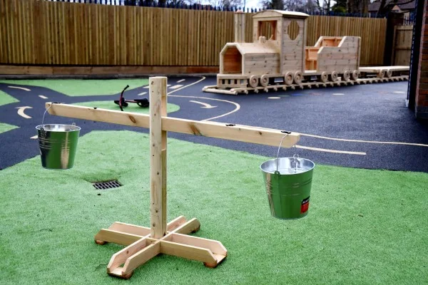 Wooden balance scale with two metal buckets, one hanging slightly lower; sits on green artificial turf in a fenced playground beside a wooden toy train and paved play area.