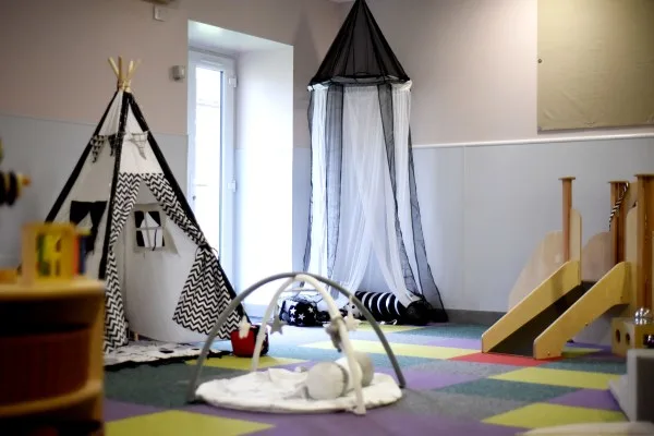 A fabric teepee stands beside a sheer hanging canopy while a baby play gym lies on multicolored foam tiles and a small wooden slide sits against pale nursery walls.