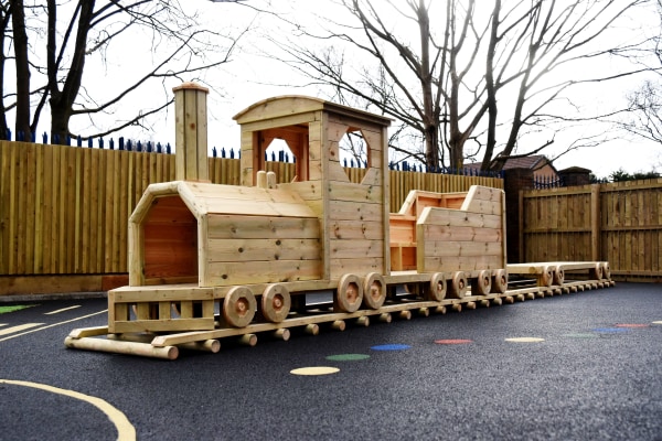 Wooden play-train sits on a black rubber playground surface, idle on its short track; surrounded by a wooden fence, leafless trees, and colored circular markings painted on the ground.