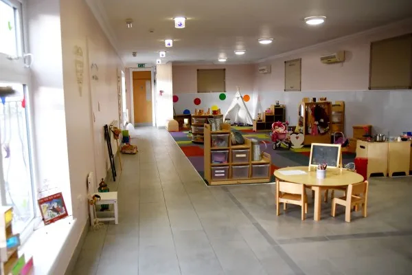 A preschool classroom with low tables and chairs, toy shelves, a play teepee and scattered toys occupies a bright tiled room with large windows and a far door marked EXIT.