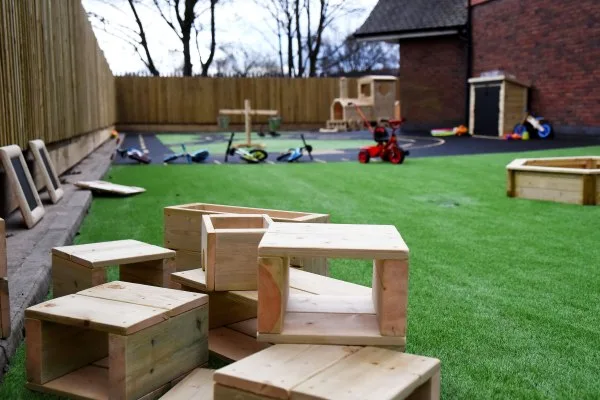 Wooden play blocks arranged and stacked on artificial grass; background shows tricycles, a red ride-on toy and a wooden playhouse near a fenced yard and brick building.