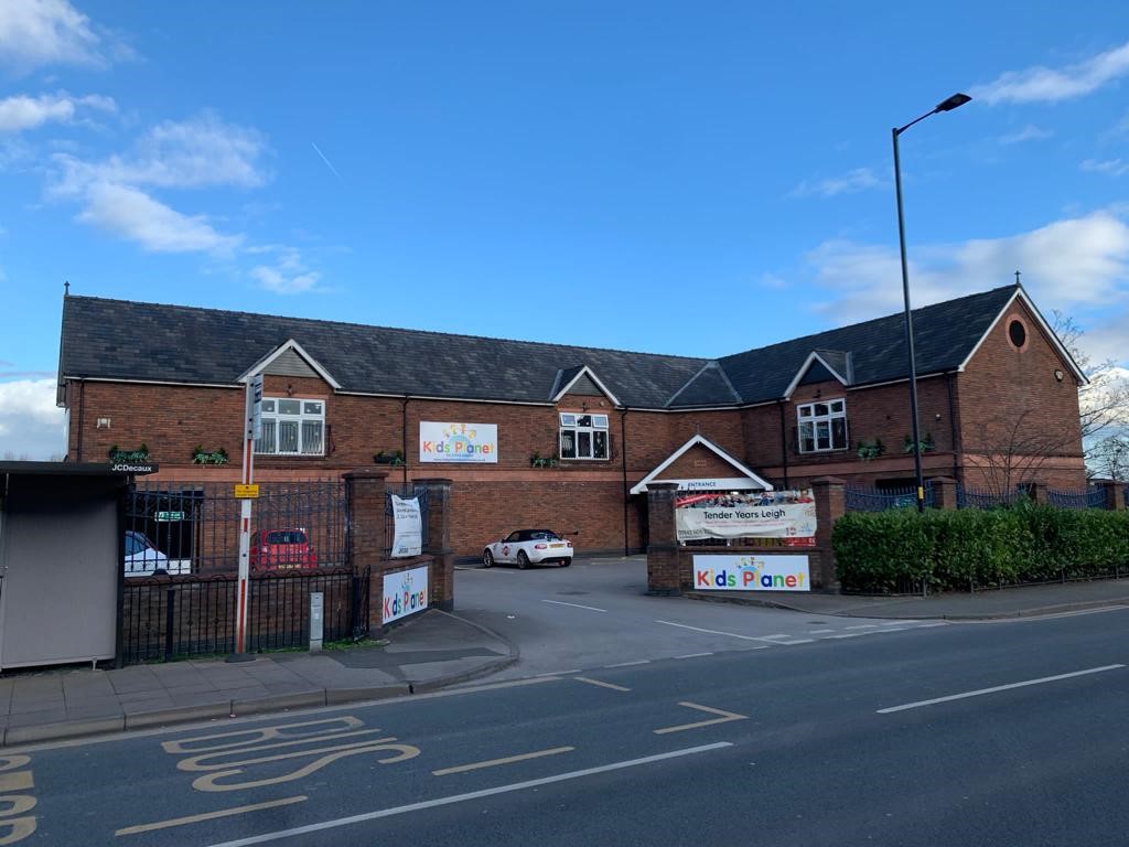 Two-story brick childcare center displays colorful signs and a canopy over its driveway; cars park by a bus stop and trimmed hedge beneath a clear blue sky.