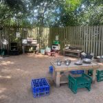 Low wooden tables and crates hold bowls, cups, and gardening tools; a small play kitchen and shelving sit against a fence; shaded leafy backyard area with bare dirt ground.