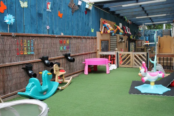 Plastic children's toys—a pink activity table, teal rocker, and unicorn rocker—sit on artificial turf, arranged under a covered, blue-paneled, decorated patio play area. Text in image: 1 2 3 4 5 6 7 8 9 10