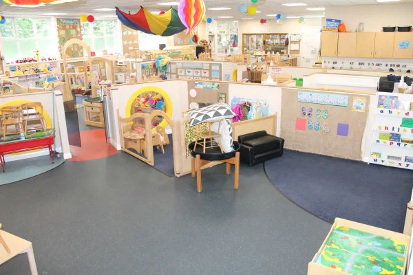 Preschool classroom — low partitions forming activity stations, toys and child-sized furniture arranged; colorful parachute and lanterns hang from ceiling; carpeted, windowed open-plan learning space. Text: a b c d e f g h i j k l m n o p q r s t u v w x y z