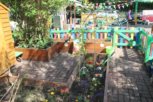 Raised wooden planters contain flowers, seedlings, a small tomato plant and scattered gardening toys; a paved courtyard surrounds them, bordered by multicolored picket fence, bunting and play dome.