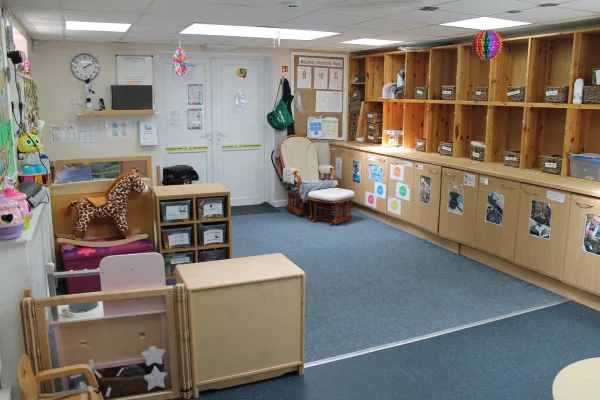 Wooden cubby shelves line a classroom wall, holding bins and toys; a rocking chair sits near double doors; low tables, a toy horse, and a carpeted play area fill the room.