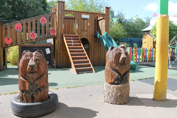 Two carved wooden bear sculptures sit atop tree-stump bases (one inside a tire) in front of a wooden playground structure with stairs and green slide, colorful picket fence and trees.