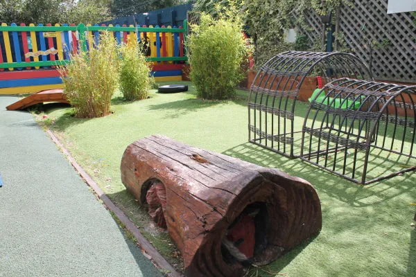 A hollow wooden log lies on artificial turf, providing a crawl-through. Nearby metal arch-climb frames stand empty; small bamboo clumps and a colorful painted fence border a children's play area.