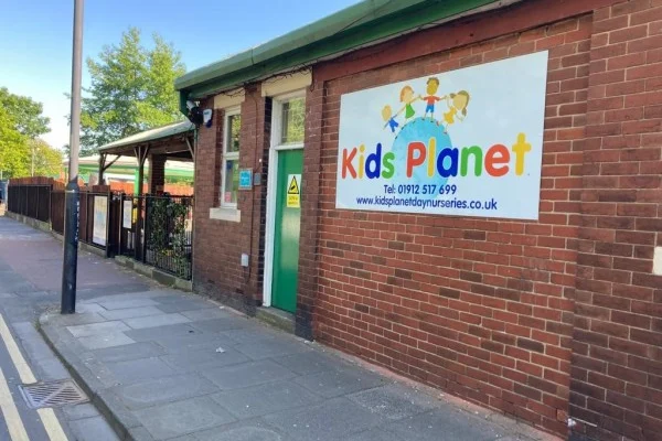 A colourful "Kids Planet" sign hangs on a red-brick nursery wall, marking the green entrance while a sidewalk and metal fence run along a quiet street.