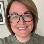 Smiling woman wearing tortoiseshell glasses; short brown hair; looking at the camera; wearing a ruffled green top; neutral wall with a framed black-and-white photo visible behind.