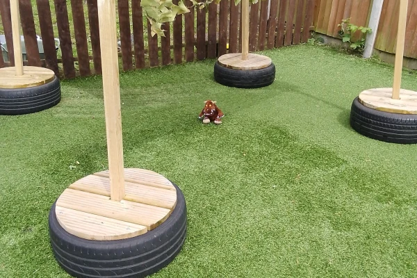 A small brown plush toy sits on green artificial turf, surrounded by wooden poles mounted in tire bases within a fenced backyard area.