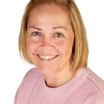 Woman smiling at the camera; short blonde hair, blue eyes, wearing a pink knit sweater; head-and-shoulders close-up against a plain white background.