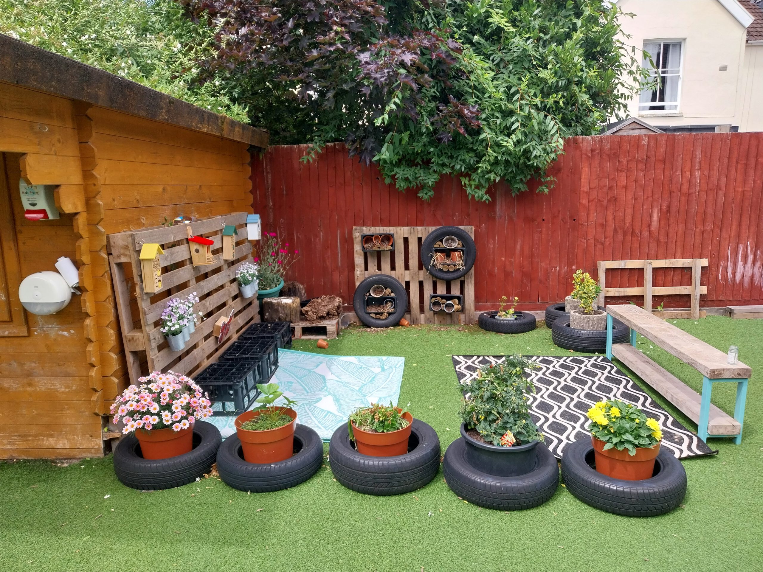 Row of five potted flowering plants placed inside recycled car tires, sitting on green artificial turf; they face a wooden shed and red fence adorned with pallets, birdhouses, and other tire planters.