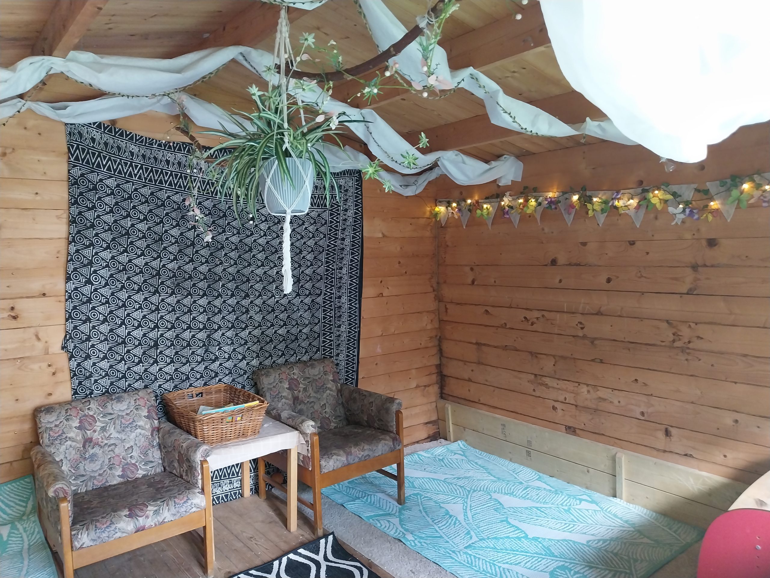 Two floral upholstered armchairs flank a small table holding a wicker basket, arranged against black-and-white patterned tapestry inside wooden shed decorated with hanging plant, draped white fabric, fairy lights.
