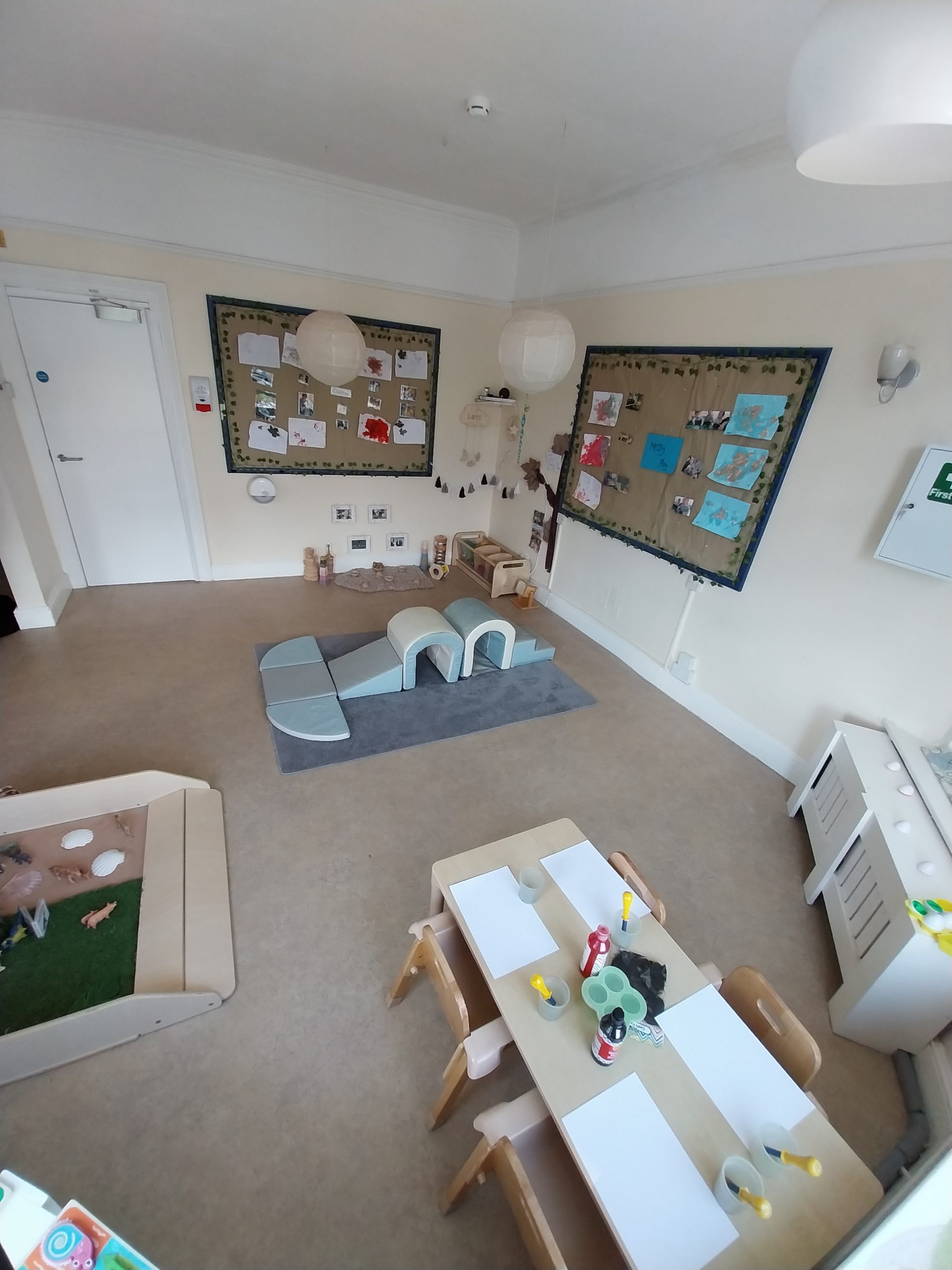 Low wooden table with blank paper, paint bottles and brushes; sits in a bright nursery with soft play arches on a rug, bulletin boards and hanging paper lanterns.