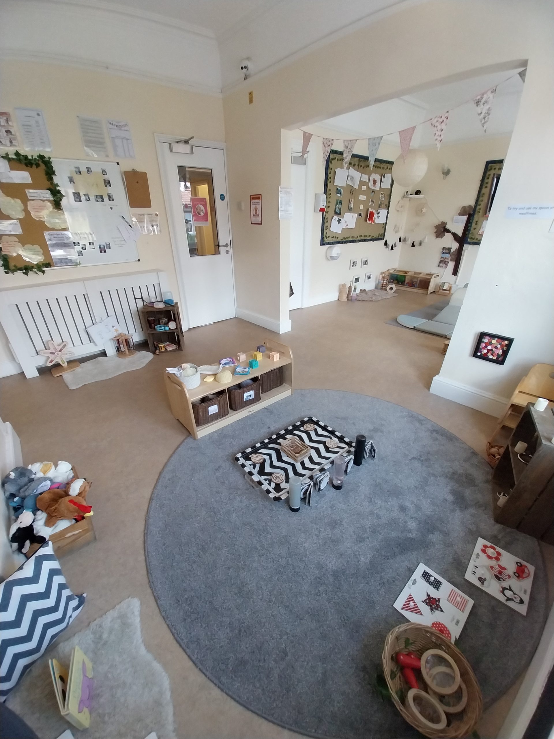 Large circular gray rug holding small wooden toys and cylinders, arranged for play; surrounded by low shelves, baskets, cushions and a bulletin board in a bright, softly decorated children's playroom.