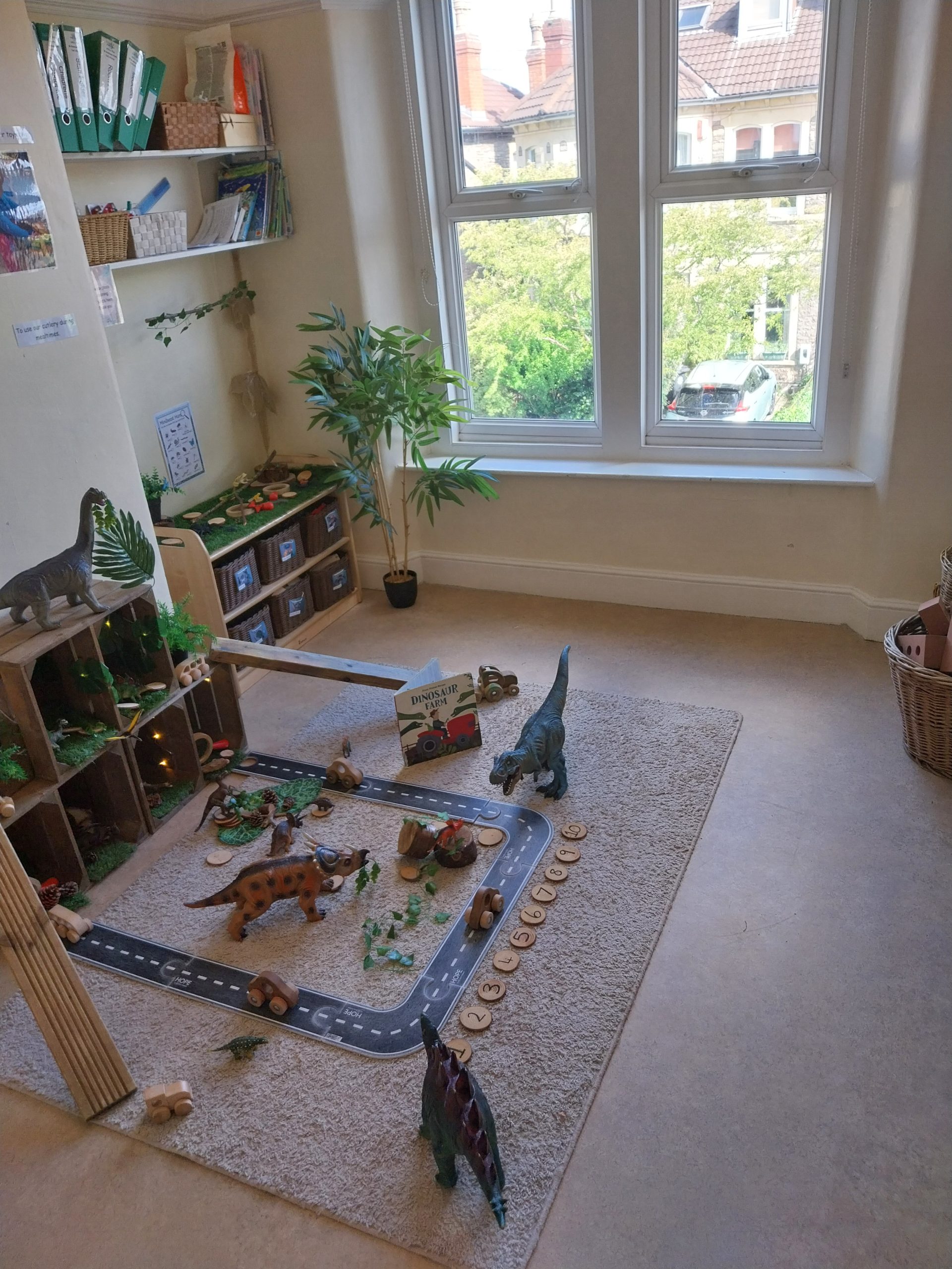 Toy dinosaurs roam a carpeted playmat, positioned around a toy road with wooden cars and nature pieces, in a sunlit corner of a children's playroom by a large window with shelves and a potted plant.
