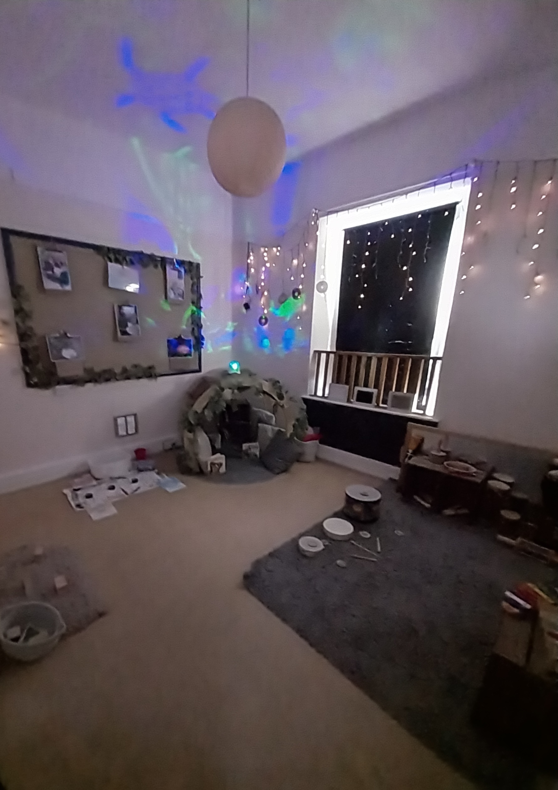 A child-sized fabric play tent glows with projected blue-green lights; fairy lights hang around a dark window; surrounding carpet, toys, bulletin board and pendant lamp fill a cozy playroom.