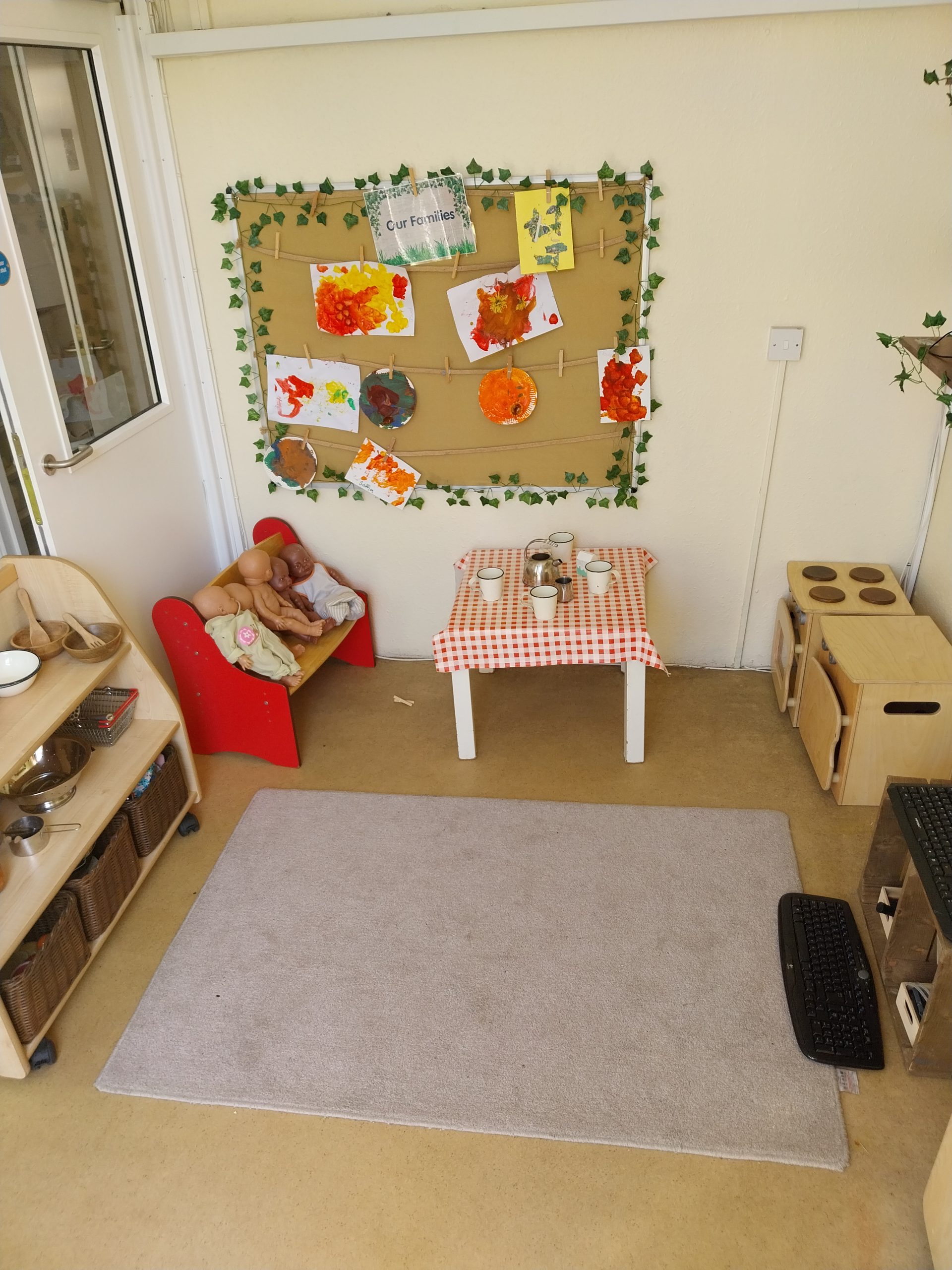 Bulletin board (text: "Our Families") displays children's painted artwork clipped to twine, in a preschool play corner with a small tea-table, dolls on a red bench, and a play kitchenette.