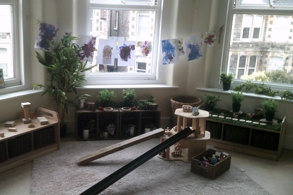 Wooden ramp with circular platform forms a toy track to a basket of small toys on a carpeted playroom floor by large windows with hanging children's paintings and potted plants.