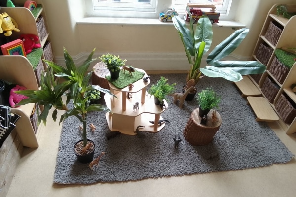 Wooden multi-level play platform holds small toy animals while potted and artificial plants encircle it on a gray rug in a sunlit children's playroom with low shelves and a window.