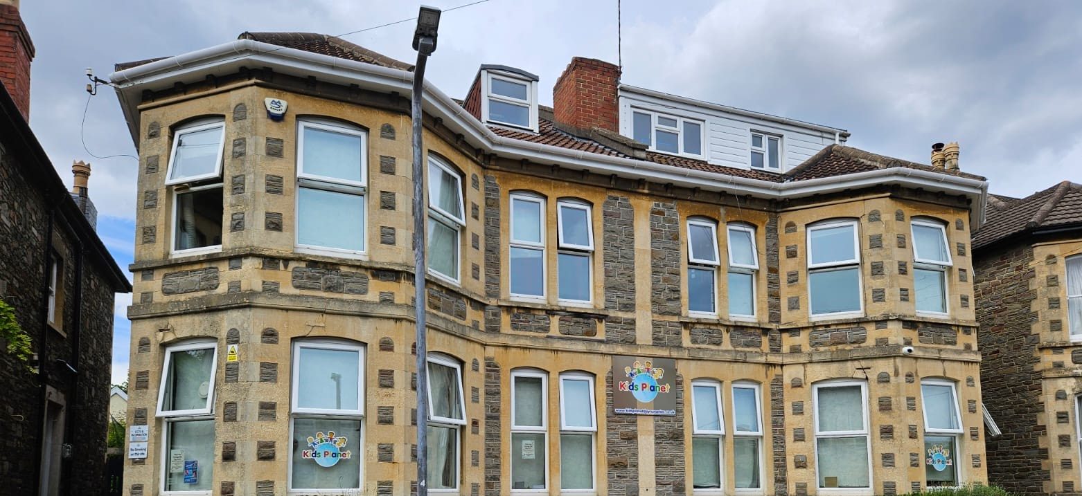 Three-story stone-and-sandstone bay building showing "Kids Planet" signage (text: "Kids Planet"), operating as a childcare center on a residential street with a lamppost and overcast sky.