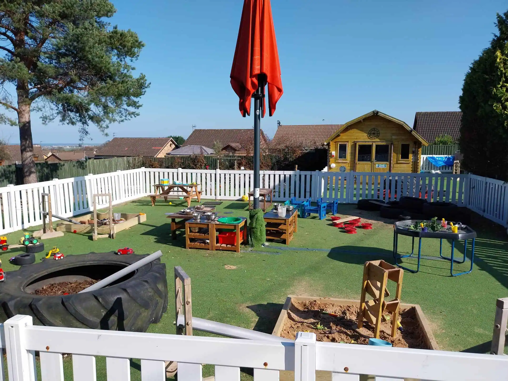 White-fenced children's play area — toy vehicles, sandboxes, activity tables and a closed red umbrella occupy artificial turf; a wooden playhouse sits at the far end with houses and trees beyond.