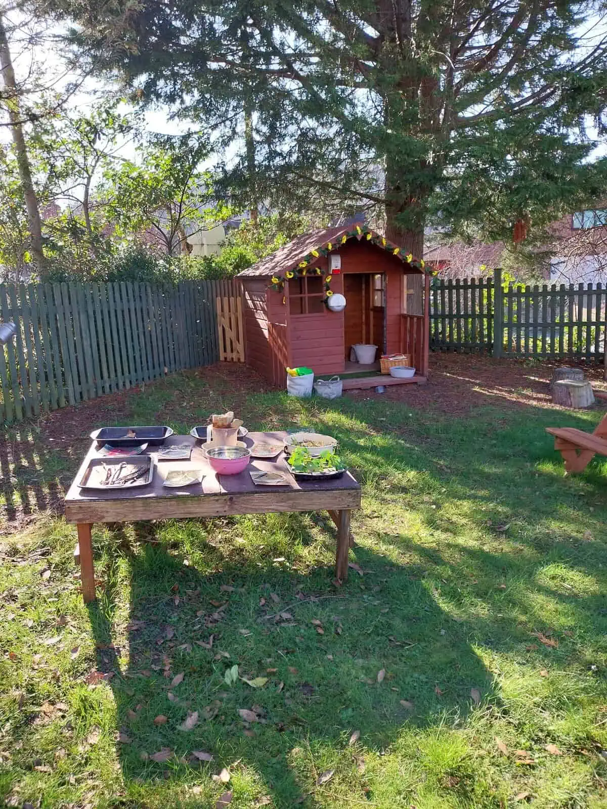 A small wooden playhouse decorated with sunflower garland stands with its door open; a low table in front displays bowls, trays, and seedlings; sunlit fenced backyard beneath tall trees.