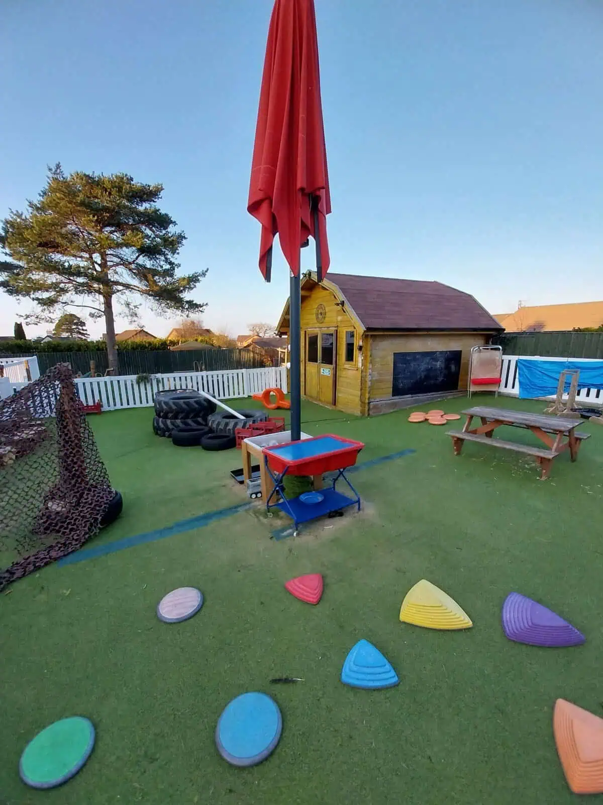 Tall closed red umbrella stands on artificial turf over a children's playground with colorful stepping stones, a water table, bench, wooden playhouse and stacked tires inside a white picket fence.