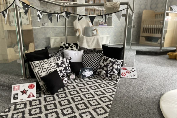 Cluster of black-and-white patterned cushions arranged in a cozy semi-circle on a geometric rug under bunting, forming a small indoor play/reading nook in a carpeted nursery area.