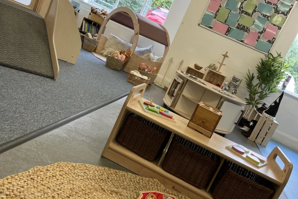 Low wooden shelf with three wicker baskets holding toys and trays, on a woven rug; nearby play kitchen, reading nook, soft play structures and windows in a bright children's classroom.