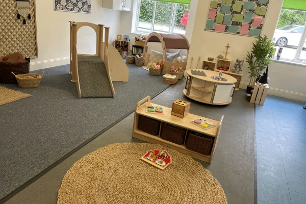 Woven round rug displays a small toy set; a low wooden shelf holds baskets and puzzles; a nearby wooden slide, play table and baskets populate a bright preschool classroom.
