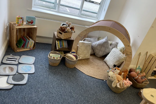 Wooden semicircular play arch cradles cushions and stuffed toys, forming a cozy reading nook on a round rug, surrounded by baskets of books and sensory mats near a window.