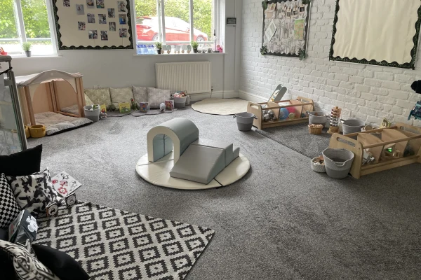 Soft foam arch and ramp placed on a round mat in a carpeted early-years classroom, surrounded by cushions, a patterned rug, low wooden toy shelves, buckets and windows.