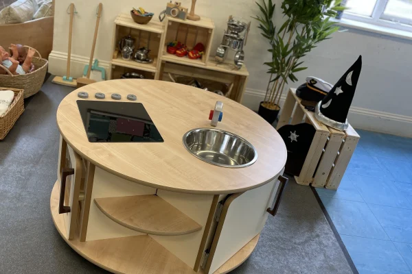 Round wooden play kitchen island holds a pretend stovetop with four knobs and metal sink; open shelves and cabinets sit amid toys, baskets, crates, a wizard hat, and a window.