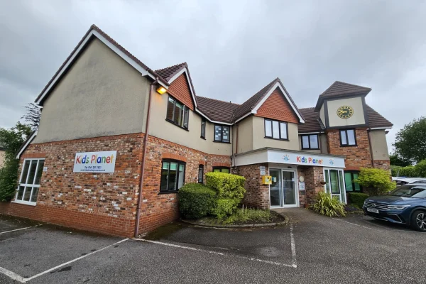Two-story brick-and-stucco childcare building bears colorful "Kids Planet" signs at the entrance and side, with shrubs, parking spaces and a parked car in front under an overcast sky.