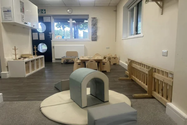A soft foam arch sits on a round padded mat; low wooden chairs, tables and storage line a bright nursery room with wood floors, a small safety gate and a back door with porthole windows.