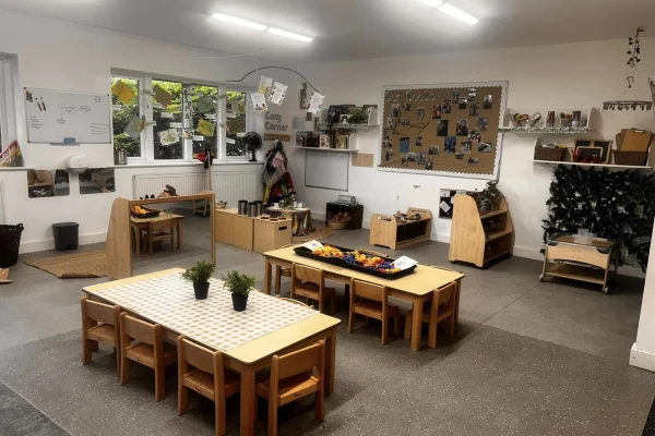Low wooden tables and child-sized chairs surround trays of craft materials and potted plants as shelving, play stations and a corkboard fill a bright, organized preschool classroom.