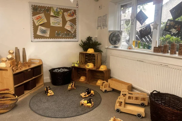 Wooden toy trucks and construction helmets sit arranged on a small round rug and stacked crates, scattered in a cozy preschool play corner with children's paintings on a bulletin board.