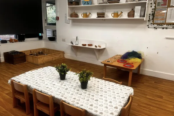 Low child-sized table draped in a patterned cloth holds two small potted plants; surrounded by wooden chairs. Classroom activity space with shelves, a sensory play table, wooden floor and a low sandpit.