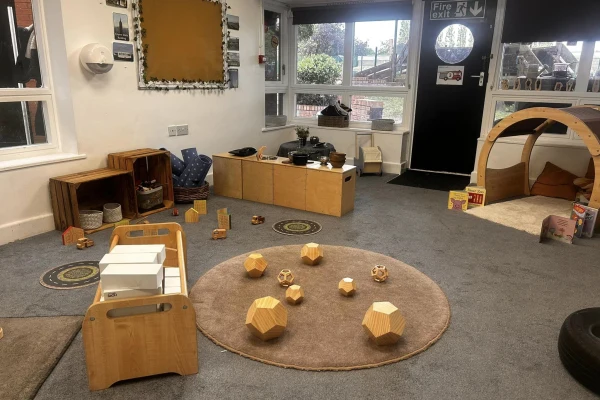 Round rug with wooden geometric blocks arranged on it; toys and low shelves nearby; wooden play arch and cubbies by windows, plus a door labeled "Fire exit" in carpeted classroom.