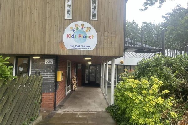 Kids Planet sign hangs above a covered entrance; a paved path leads into a shaded corridor between brick walls, flanked by wooden fencing and dense green shrubs.