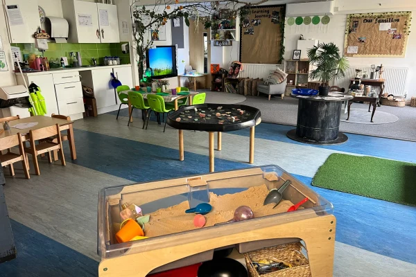 Wooden sand table (foreground) holds scoops, shovels and toys, sitting idle; a child-sized classroom surrounds it with low tables and green chairs, activity stations, kitchenette and carpeted reading corner.