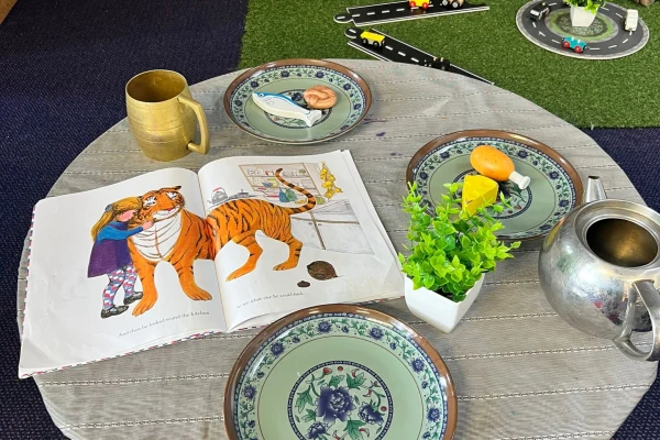 A children's picture book lies open, showing a girl hugging a tiger; it rests on a round table with three decorative plates, a brass mug, a small potted plant, and a metal teapot. Text on the book's pages is too small or blurred to transcribe.